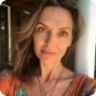 A woman with wavy hair is standing outside, wearing a patterned orange top. She is looking directly at the camera with a neutral expression. The background is slightly blurred, featuring a part of a building and a column.