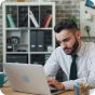 A man with a beard sits at a desk, focused on a laptop. He wears a white shirt and green tie. The background features office shelves with books and decor against a brick wall. Pens and a notepad are on the desk.